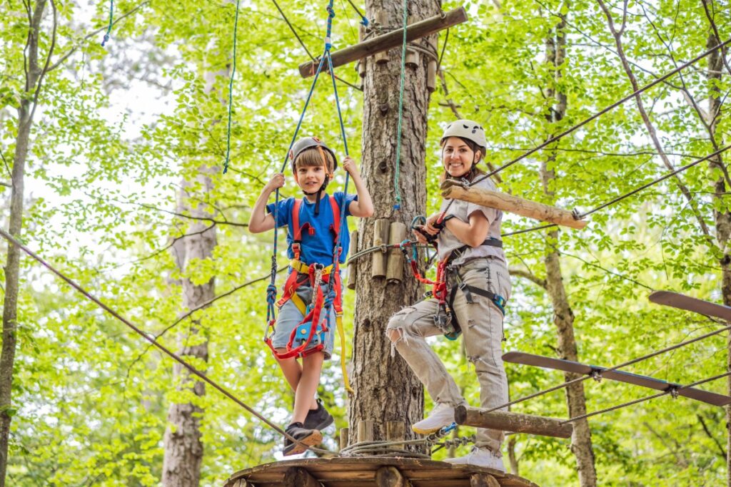 Mother and son climbing in extreme road trolley zipline in forest on carabiner safety link on tree to tree top rope adventure park. Family weekend children kids activities concept Portrait of a
